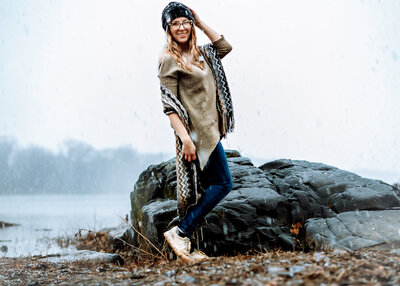 A woman is standin with one hand on her head smiling. There's trees foggily in the background, water, and large rocks right behind her and it's snowing.