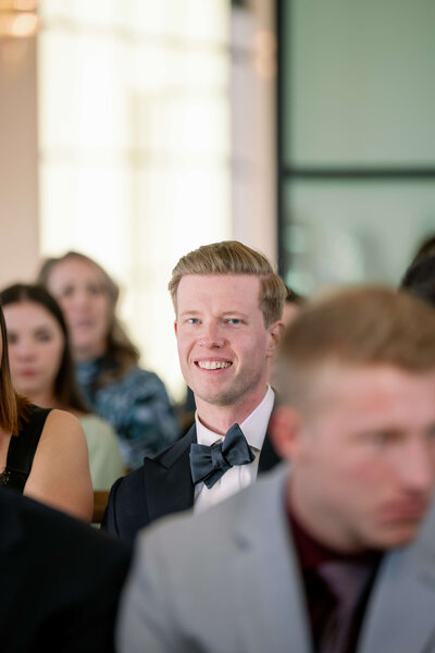 Image of a groomsman sitting, looking at the bride and groom smiling