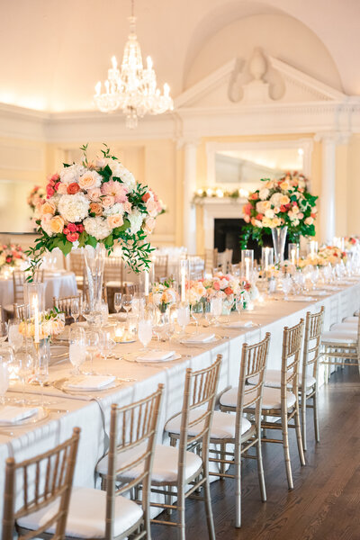 An elegant wedding reception setup with long tables adorned with tall flower arrangements of pink and white roses. Gold chairs are neatly arranged around the tables. Chandeliers hang from the ceiling, creating a warm and inviting atmosphere.