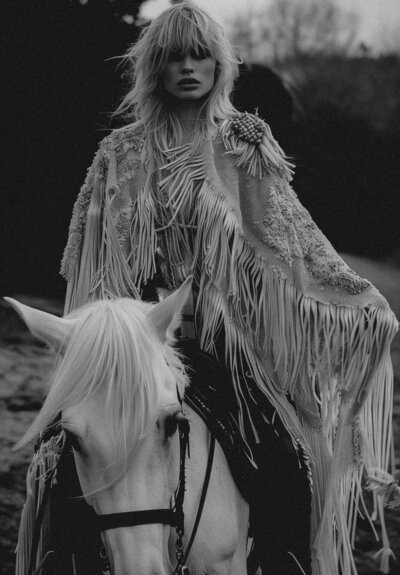 A person with long, tousled hair and a fringed cloak sits on a white horse outdoors. The black-and-white image captures the spirit of Sweet Pea Ink Creative, blending artistry and inspiration ideal for brand design.