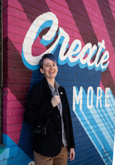 photographer River Dantzler smiles and leans against a painted wall with  words "create more". they have short blue hair and wear a blazer with a rainbow pride pin and "they/them" pronoun pin