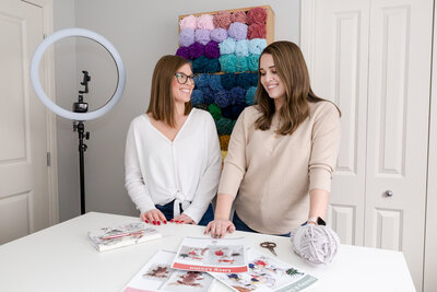 Calgary brand photography shot of two sisters who are smiling at one another. They are in their Double Dutch Crochet Co design and craft room, with a ring light and yarn behind them on the wall.