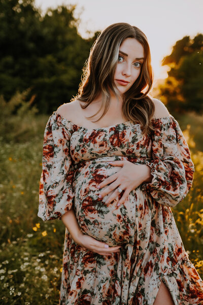 A expecting mom holding her belly in a floral dress and looking at the camera at Northwest Community Park in Fort Worth, Texas
