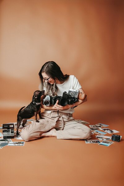 A destination wedding photographer sits cross-legged on a warm-toned studio backdrop, holding multiple vintage and modern cameras while smiling at a small dog. Surrounded by printed photos and old-school camera gear, she exudes a playful, nostalgic vibe.