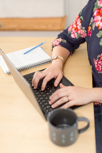 Woman's hands on a black laptop next to a grey coffee mug