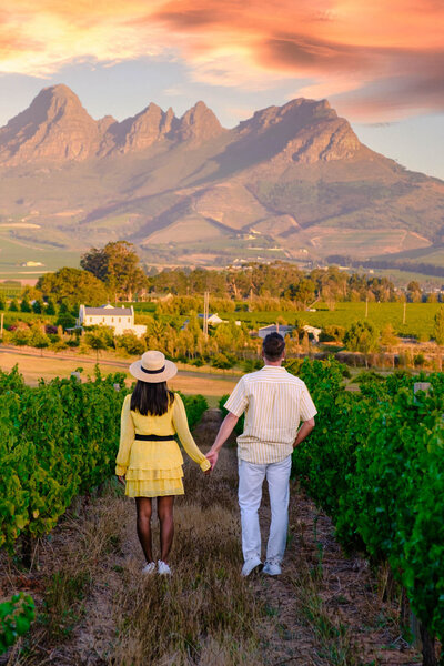 A couple holding hands walks through a vineyard at sunset, with a mountainous landscape in the background. The sky is a vibrant mix of orange and pink hues. The woman wears a yellow dress and hat, and the man wears a striped shirt and white pants.