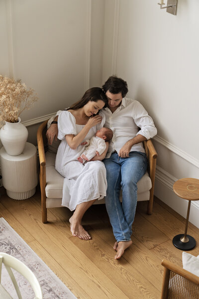 A couple sit close in a loveseat with their baby boy sleeping between them during a natural newborn photo shoot with Jess Morgan Photography at their home in London's Kensington.