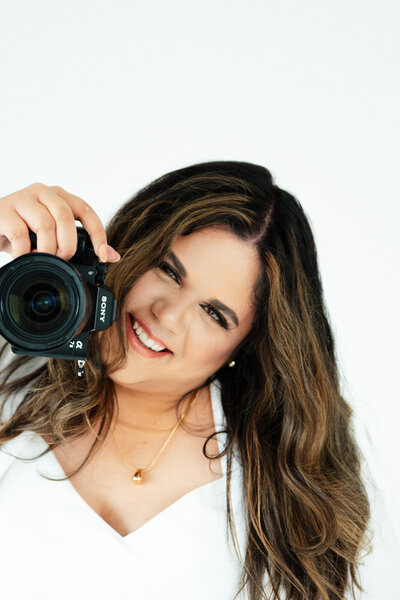 Adriana Montoya of Adriana Montoya Photo is holding her camera in her right hand with it pointing towards the camera. She is smiling with her head slightly tilted to her left.