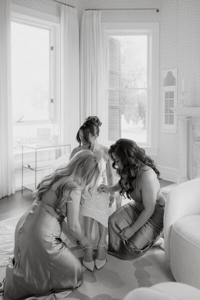 Black and white photo of bridesmaids kneeling down helping bride put on her shoes