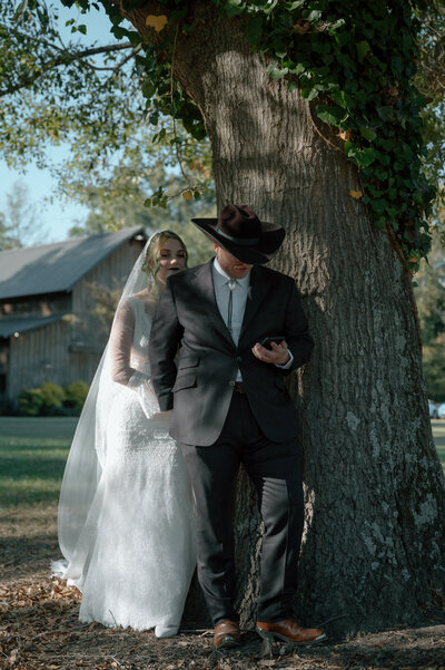 a bride and groom stand against a tree in western wedding attire
