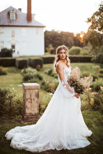 Bride in lace wedding gown holding bouquet in the garden at sunset outside a historic Portland Maine estate.