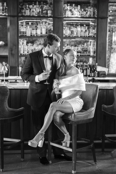 A couple dressed elegantly in formal attire enjoy drinks at a bar. The man in a tuxedo stands beside the woman, who is seated on a stool wearing a sleek off-shoulder dress and high heels. Shelves of bottles fill the background. Black and white photo.