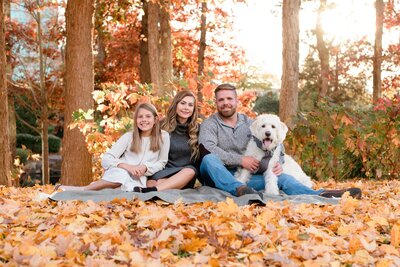 Family and their dog sit on a blanket surrounded by autumn leaves in a wooded area.