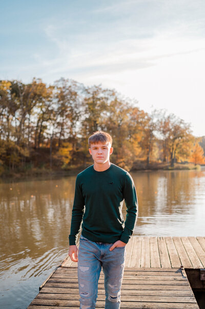 A senior boy walks on a lake dock and smiles at the camera.
