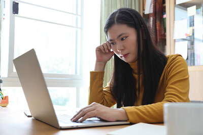 An Asian woman assistant professor sits at a desk staring at a laptop with a concerned expression. She is leaning lightly on one hand and using the laptop tracker pad with the other.