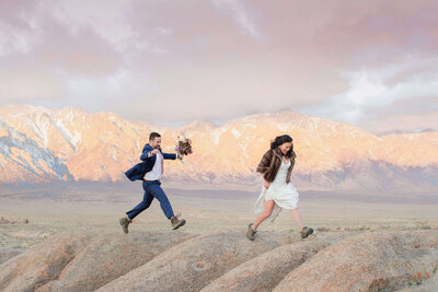 A bride and groom leap across large boulders at Alabama Hills, in front Mt Whitney in the high california desert. 