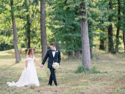bride and groom walk together with field with tall trees behind them in Arkansas