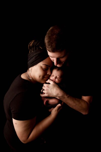 A high-key family photo from a newborn session by Mobile AL Newborn Photographer. The newborn baby boy is being held by his dad, safely resting on dad's chest and tucked under his chin. Mom comes in from the left side to rest her face against the face of her baby, as she helps dad craddle the baby with her hand under the baby's feet. Everyone is wearing black, and are on a black backdrop for an intimate portrait.
