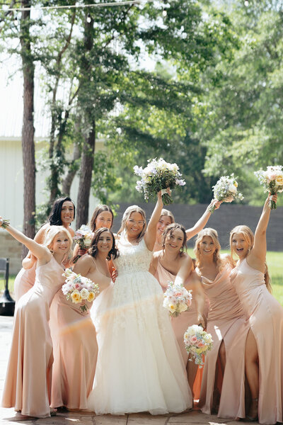 Bride and bridesmaids in pink dresses holding up bouquets in a meadow