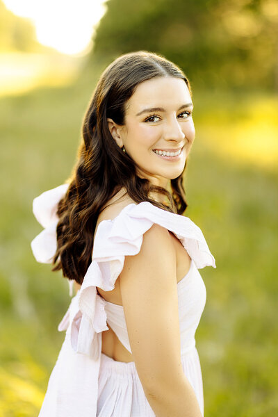 A woman with long dark hair smiles at the camera, wearing a light, ruffled dress. She stands in a sunlit outdoor setting with greenery in the background, perfectly captured by a Brainerd Lakes Area Photographer.