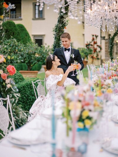 A couple poses in an enchanting outdoor wedding setting at a historic Italian venue, with chandeliers, vibrant flowers, and a stunning backdrop. The bride wears a Monique Lhuillier gown, and the groom is in a sharp suit, captured by Tuscany wedding photographer Andreas K. Georgiou.