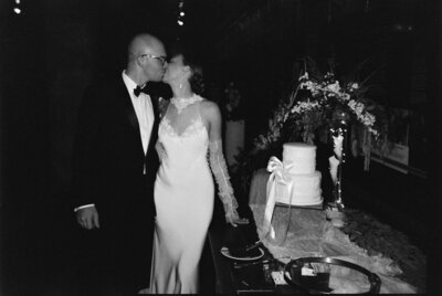 black and white image of bride and groom kissing on their wedding day standing beside cake on table photo by tom thornton photography of minnesota