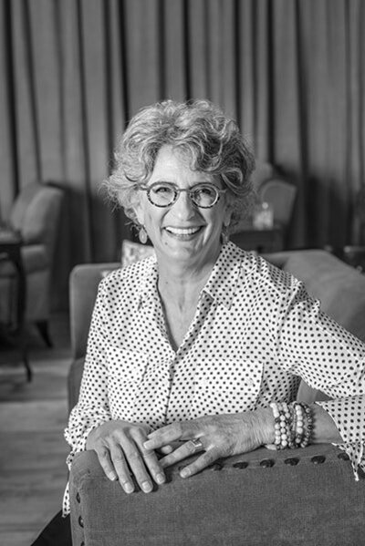 Amy Posner smiling wearing glasses and a polka dot shirt, resting her arm on a chair in a monochromatic setting.
