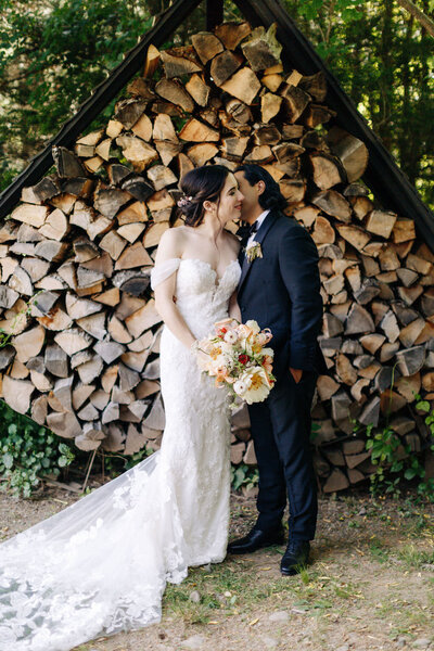 Bride in lacy white dress with romantic train leans into groom as he whispers in her ear, in front of chopped wood and forest at Foxfire Mountain