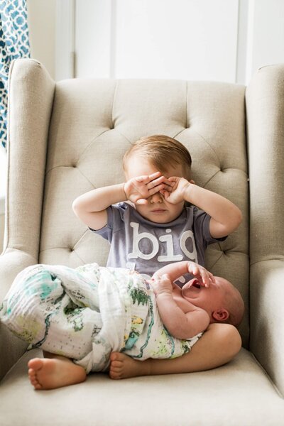 Toddler covering eyes while sitting with crying baby on a beige armchair during a newborn lifestyle photography session.
