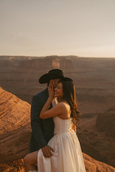 A couple embracing and smiling while looking opposite directions in their wedding attire at sunset in utah with canyon views in the background