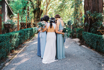 Bride and Bridesmaids huddle together before the wedding ceremony. Bridesmaids are wearing mismatched dusty blue toned chiffon dresses