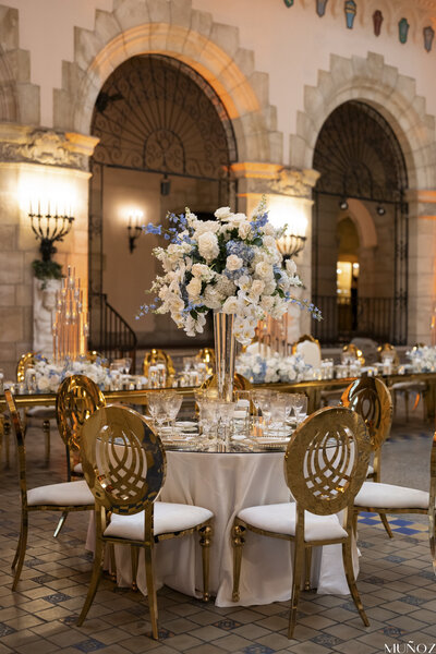 A beautifully set wedding table with a tall floral centerpiece and gold chairs, capturing the luxurious decor style of Socially Significant Events.