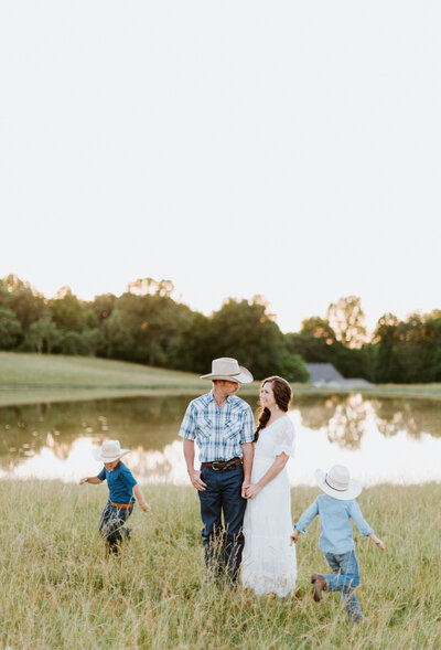 Family photo of wedding photographer in Nashville with husband and kids in cowboy hats on the farm by a pond