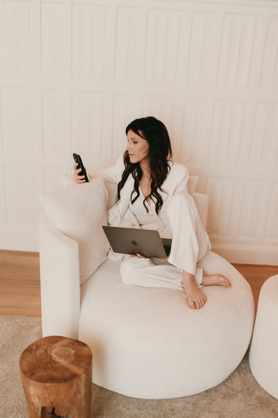 Vrouw dat zit in een zetel met gsm in de hand en laptop op de schoot