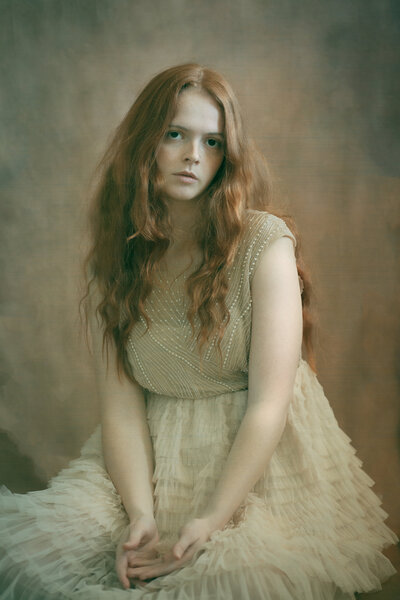 A girl with long red hair wears a white tulle dress sitting gracefully on the ground in front of an antique backdrop