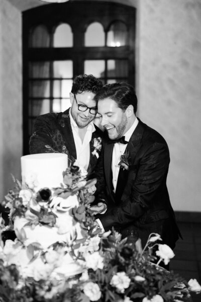 Two Grooms cut their cake in black and white with a pile of flowers filling the foreground.