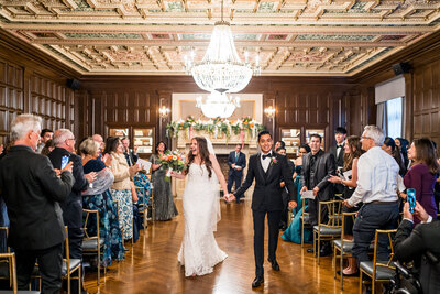 Stella and Jair walk back down the aisle during their wedding recessional at the Athletic Club of Columbus, beaming with joy and big smiles as guests cheer them on.