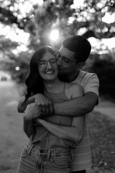 A couple sharing a joyful moment in Spring City Park. The man kisses the woman's cheek as they both smile, surrounded by trees.