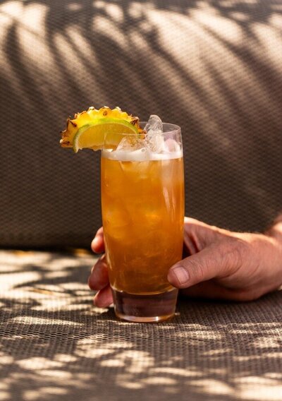 Tropical cocktail with ice, garnished with lime, orange, and pineapple peel, held by a person resting on a textured surface in dappled sunlight.