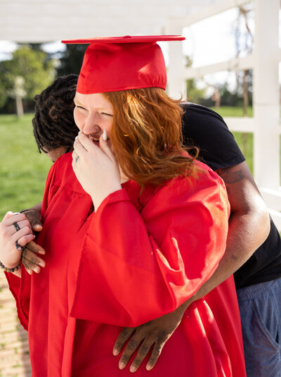 Natalie Dew, a 2021 graduate, laughing with her boyfriend and wearing her red cap and gown. This photo was take at Slate Farm Living Historical Farm in Canal Winchester, Ohio.