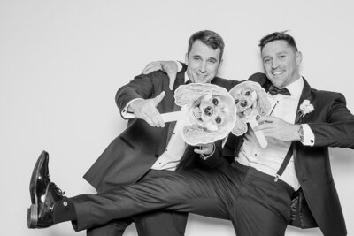 Two groomsmen in tuxedos posing in a black and white glam photo booth, holding oversized cut-out props of dogs for a fun and personalized wedding touch, captured by Philly Snap Booth.