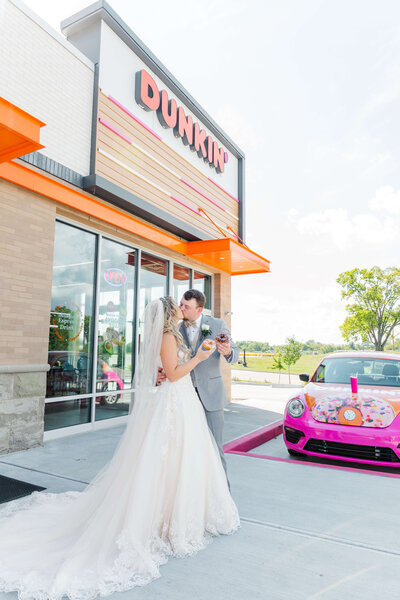 Bride and groom kiss each other in front of a dunkin donuts holding donuts