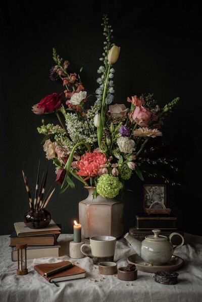 Stilleven met bloemstuk in zandkleurige vaas van aardewerk met rondom vintage boeken, servies en accessoires.