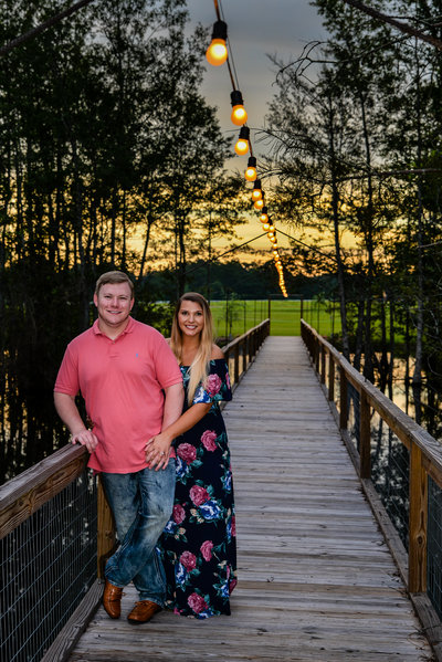 Engaged couple on a wooden bridge with Edison lights above them with a pretty sunset behind them by Allison Burton Photography