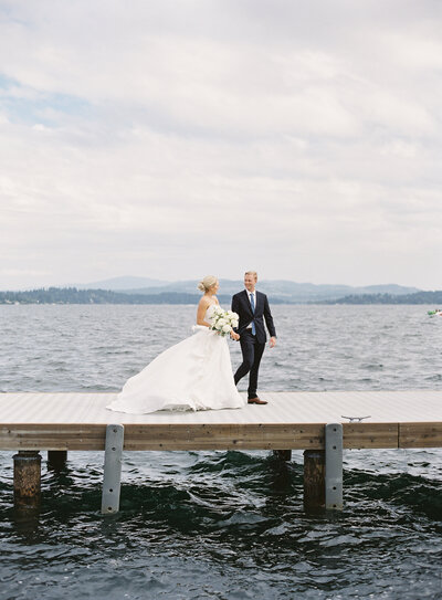 bride and groom walking on dock holding hands over lake water with wind blowing