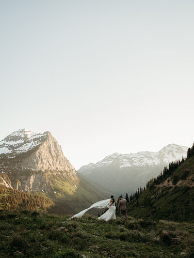 couple standing in front of the grand mountains of glacier national park with snow on them