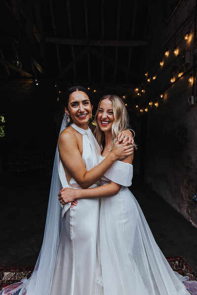 Two stylish brides smiling and hugging at their wedding at The Cowshed Wood at Hall Farm, with a dark backdrop and twinkling festoon lights creating a magical atmosphere