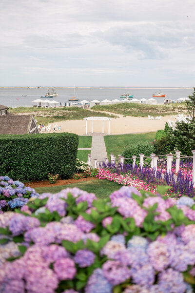 A vibrant garden filled with purple hydrangeas and colorful flowers leads down a manicured path to the beach at Chatham Bars Inn, with sailboats and beach chairs dotting the coastal horizon.