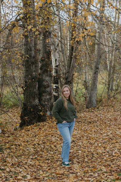 A woman standing next to trees wearing a green sweater and blue jeans.