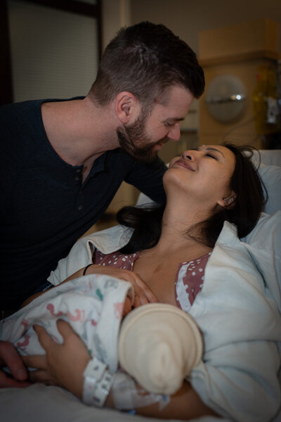 a husband leans to kiss his wife while she is laying in a hospital holding her newly born baby at UW Medical Center at Montlake in Seattle, WA.